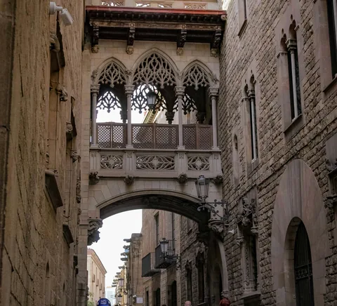 People walking below the Gothic Quarter bridge between stone buildings.