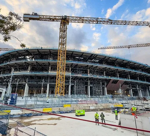 Camp Nou exterior under redevelopment, cranes above the modern stadium tiers, with small construction workers in the foreground; photographed 12 December 2025.