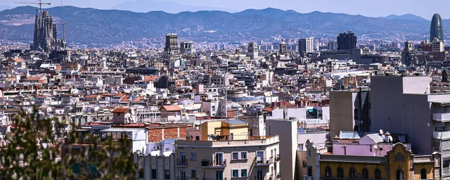Daylight view across Barcelona rooftops toward Sagrada Família and distant hills.