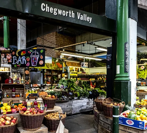 Actual Borough Market produce stall, photographed January2020: baskets of fruit/vegetables, iron market structure and modest human background activity.